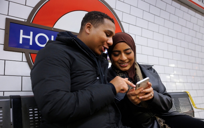 Two people sit on a bench underground looking at a phone